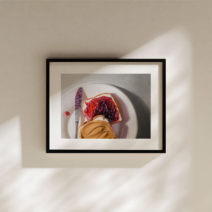 Fine art print of a peanut butter and jelly sandwich on a plate, framed in black with a white mat, hanging on a wall.