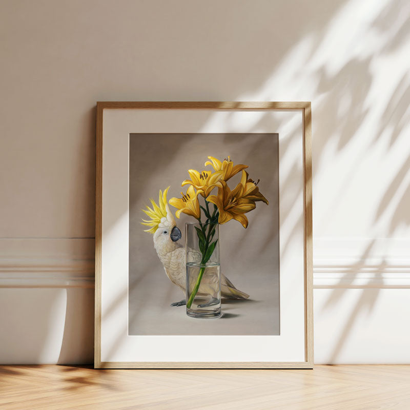 Fine art print of a white sulphur-crested cockatoo and yellow lilies in a glass vase, framed in a light wood frame.