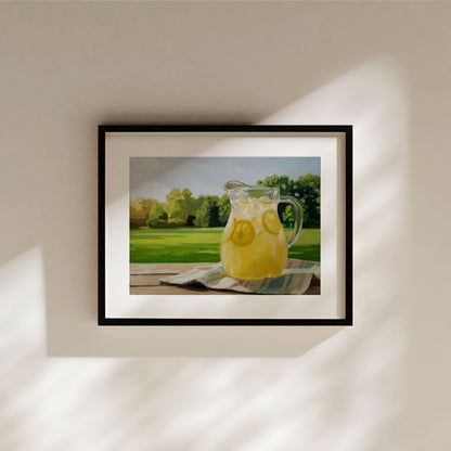 Framed fine art print of a lemonade pitcher on a wooden table, set against a sunny, vibrant green landscape.