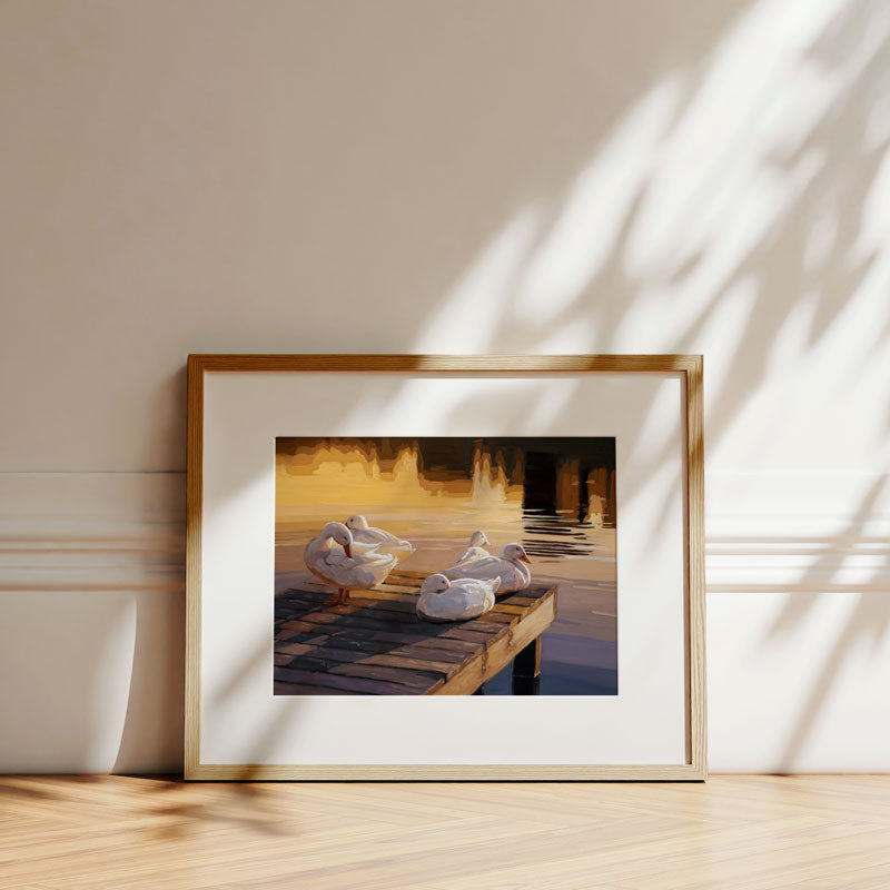 Framed fine art print of white ducks on a wooden dock at sunset, displayed in a light wood frame against a neutral wall.