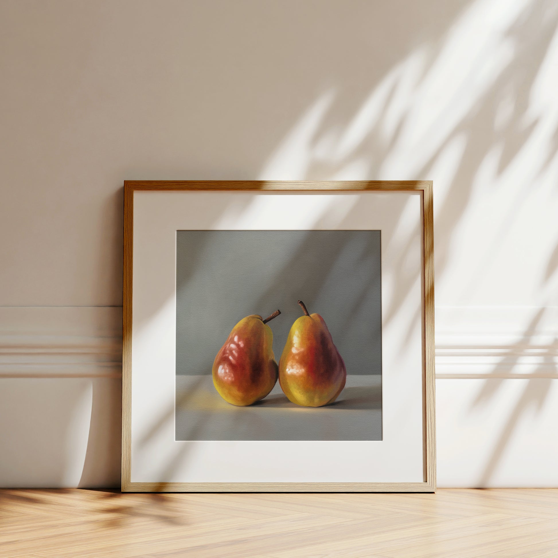 Fine art print of two ripe, red and yellow pears in a light wood frame, leaning against a white wall.