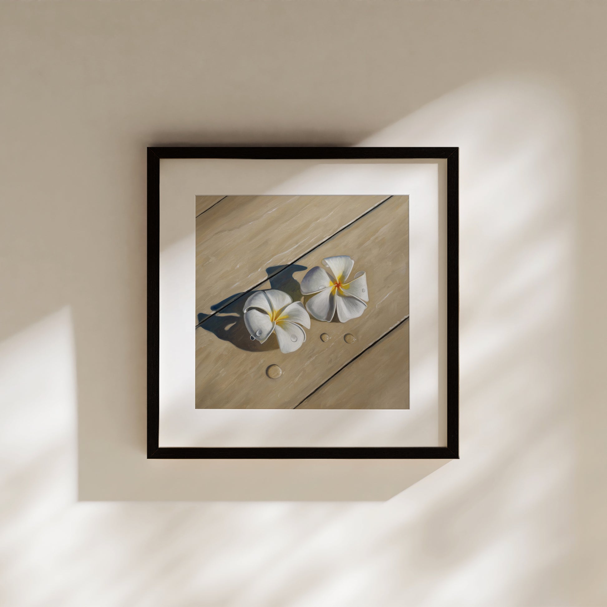 Framed fine art print of two white plumeria flowers with water droplets on light wood, hanging on a cream wall.
