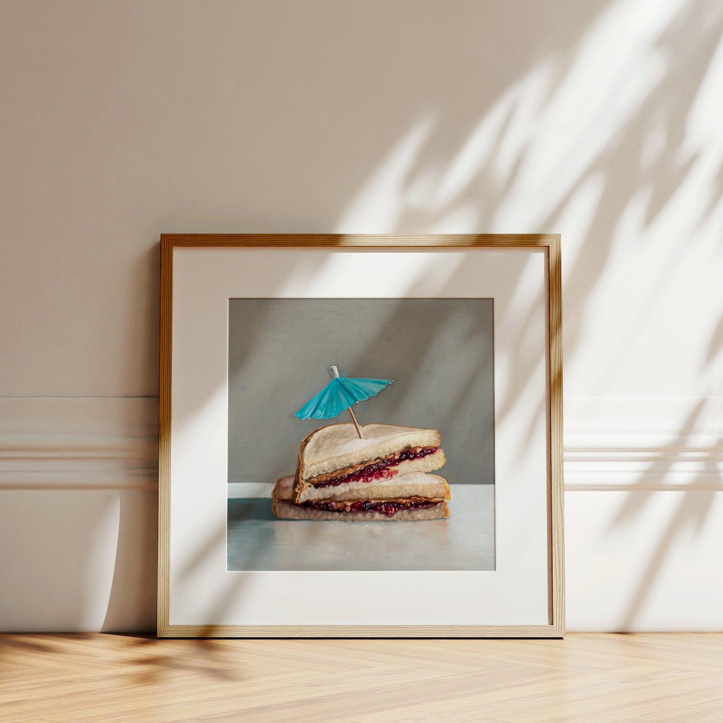 Framed fine art print of a peanut butter and jelly sandwich with a blue cocktail umbrella, on a light wood floor.