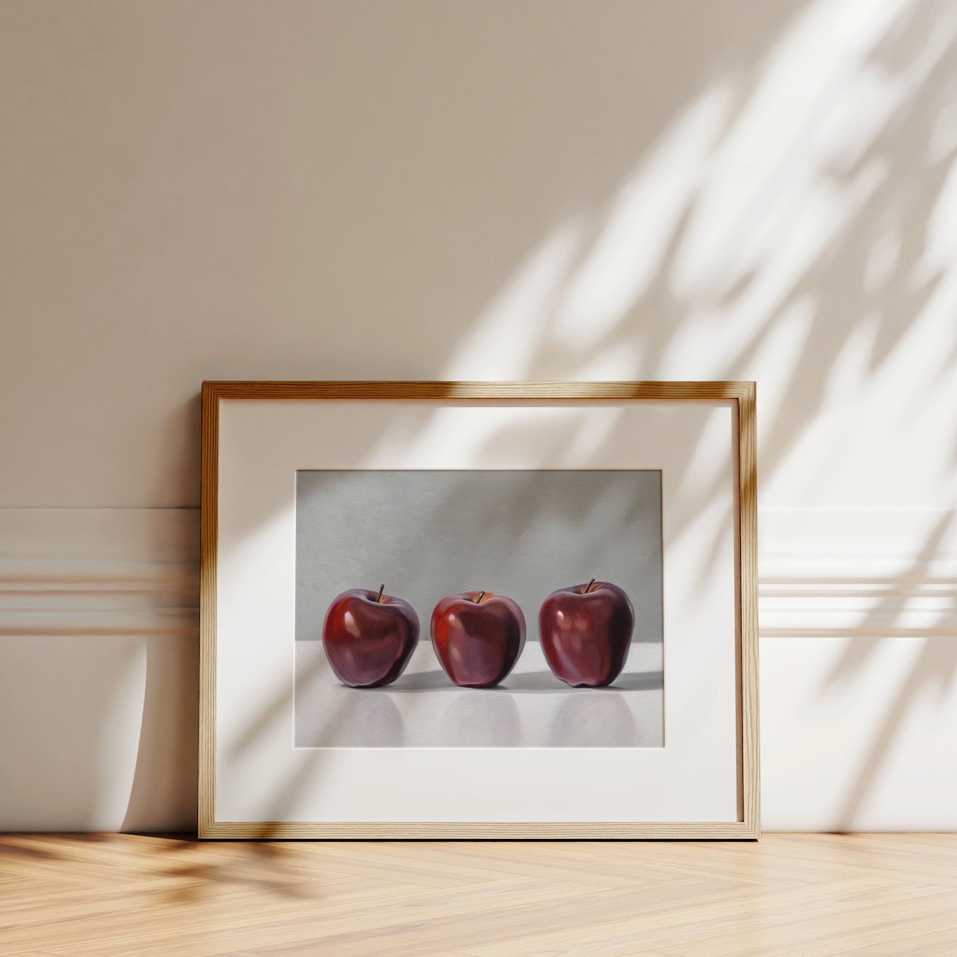 Framed fine art print of three vibrant red apples on a white surface, with natural light and shadows.