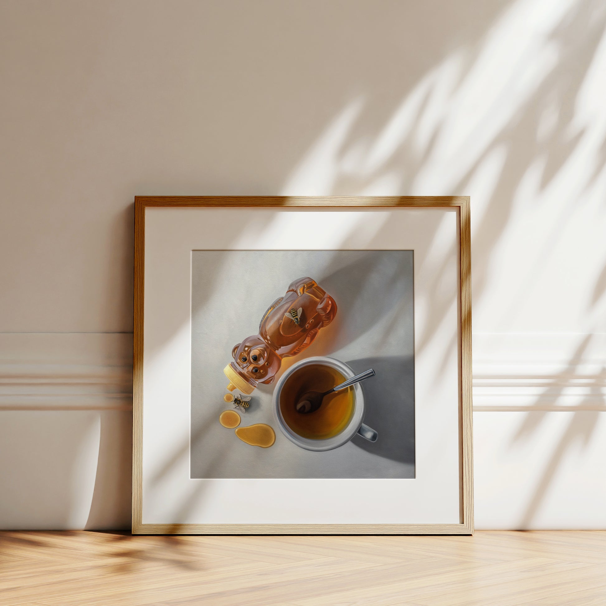 Fine art print of a honey bear bottle, spilled honey, two bees, and a cup of tea with a spoon, framed in light wood.