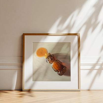 Fine art print of a honey bear bottle spilling golden honey, framed in light wood, leaning on a wall.