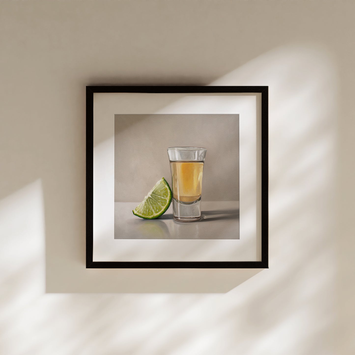 Framed fine art print of a tequila shot and a vibrant green lime wedge, hanging on a warm beige wall.