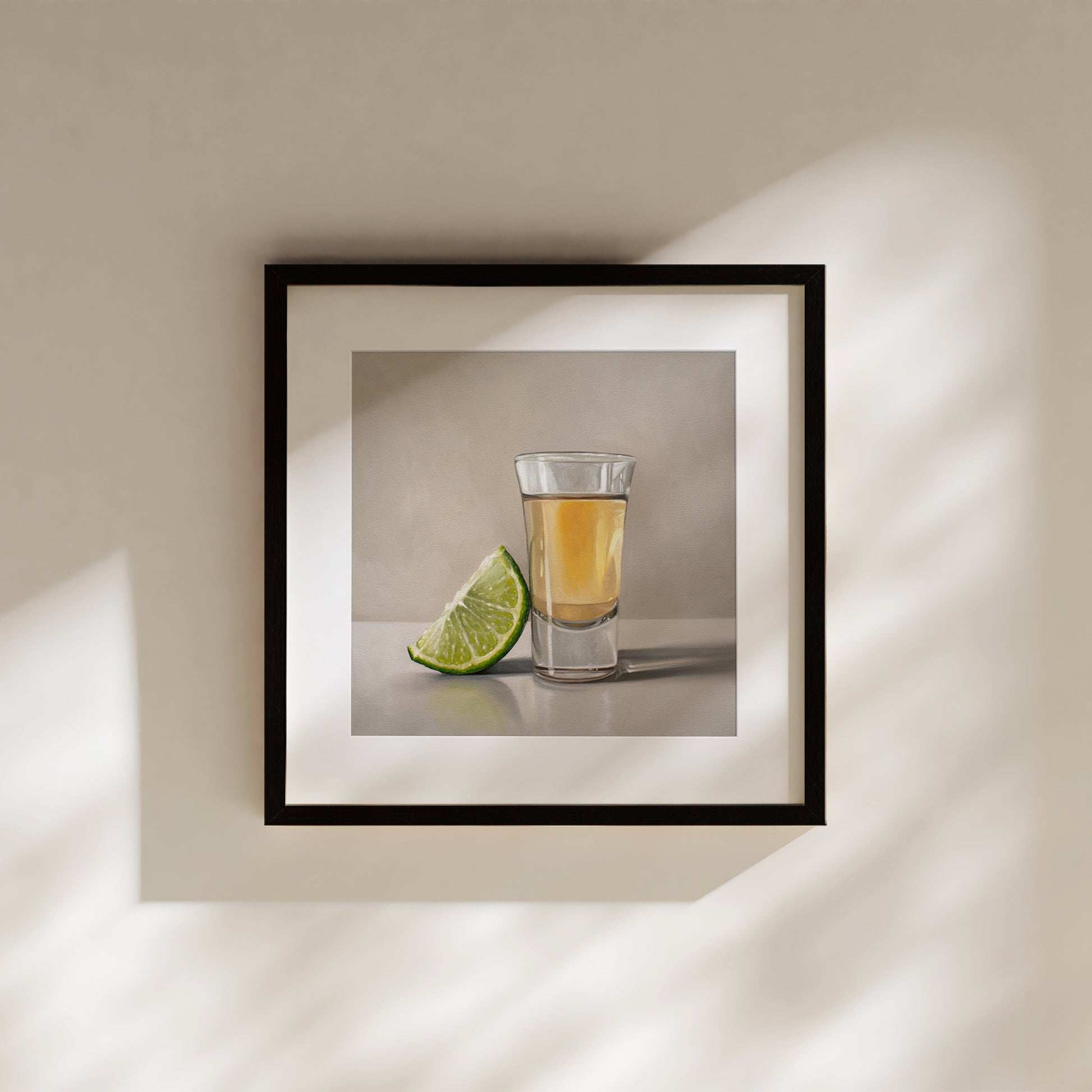 Framed fine art print of a tequila shot and a vibrant green lime wedge, hanging on a warm beige wall.