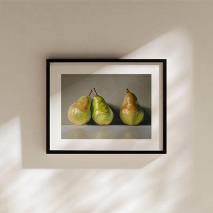 Framed fine art print of three green and red pears on a grey background, hanging on a cream wall.