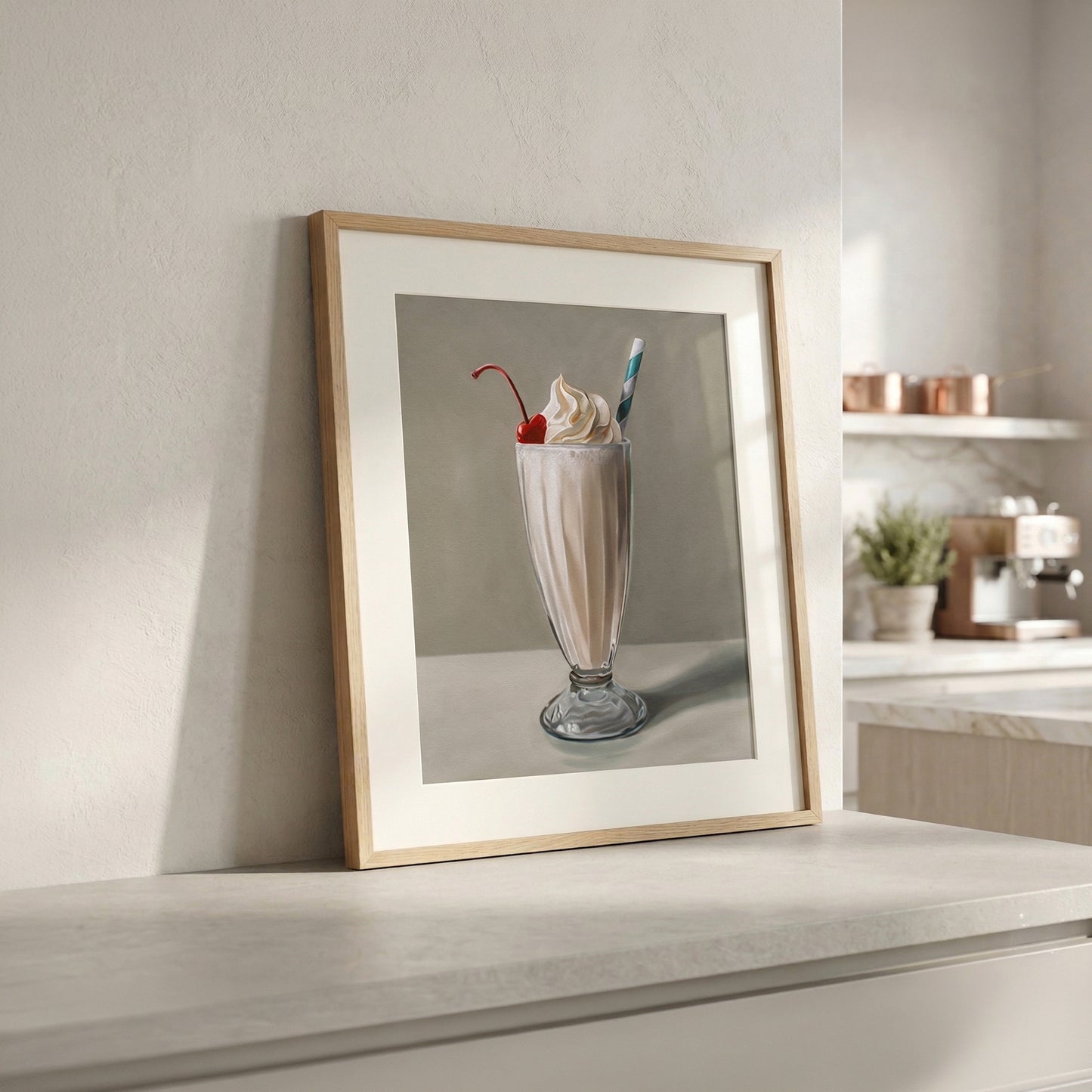 Framed fine art print of a vanilla milkshake with whipped cream and a cherry, leaning against a white wall in a modern kitchen.
