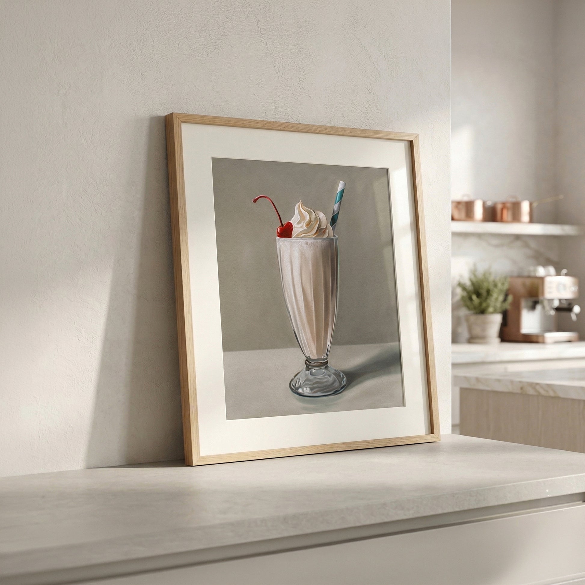 Framed fine art print of a vanilla milkshake with whipped cream and a cherry, leaning against a white wall in a modern kitchen.
