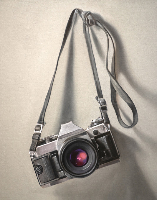 Fine art print of a vintage silver and black film camera with a strap, hanging against a light background.